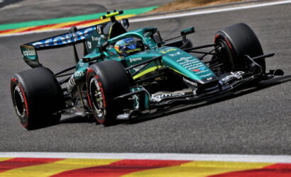 Formula 1 race car in teal and black livery speeding around a track, driver in helmet visible, on a curving corner with colorful run-off paint behind.