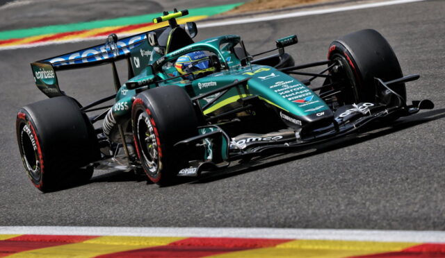 Formula 1 race car in teal and black livery speeding around a track, driver in helmet visible, on a curving corner with colorful run-off paint behind.