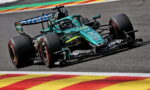 Teal Formula 1 car rounds a corner on a race track, with colorful curbs in the foreground and sponsor decals visible on the body.