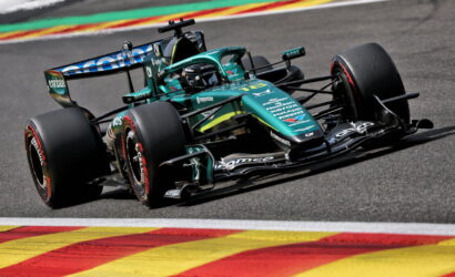 Teal Formula 1 car rounds a corner on a race track, with colorful curbs in the foreground and sponsor decals visible on the body.