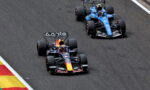 Two Formula 1 cars race side by side on a track, a Red Bull car ahead of a blue car near a left curb.