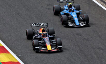 Two Formula 1 cars race side by side on a track, a Red Bull car ahead of a blue car near a left curb.