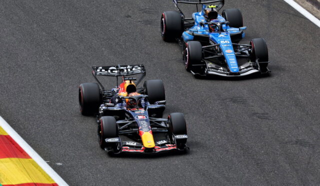 Two Formula 1 cars race side by side on a track, a Red Bull car ahead of a blue car near a left curb.