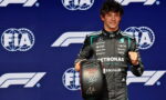 F1 driver on a podium holding a signed Belgium 2026 tire trophy, FIA logos in the background.