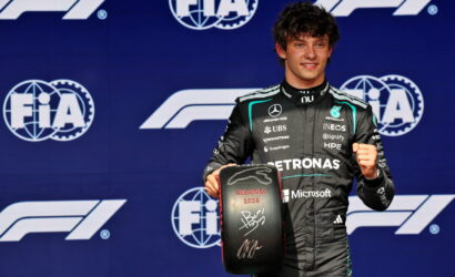 F1 driver on a podium holding a signed Belgium 2026 tire trophy, FIA logos in the background.