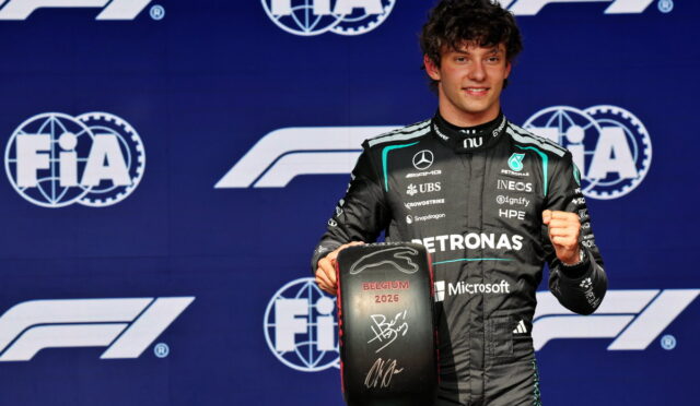 F1 driver on a podium holding a signed Belgium 2026 tire trophy, FIA logos in the background.