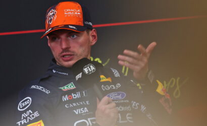 Male race car driver wearing an orange and black Oracle Red Bull Racing cap giving a press conference, speaking into multiple microphones with team logos visible on his suit.