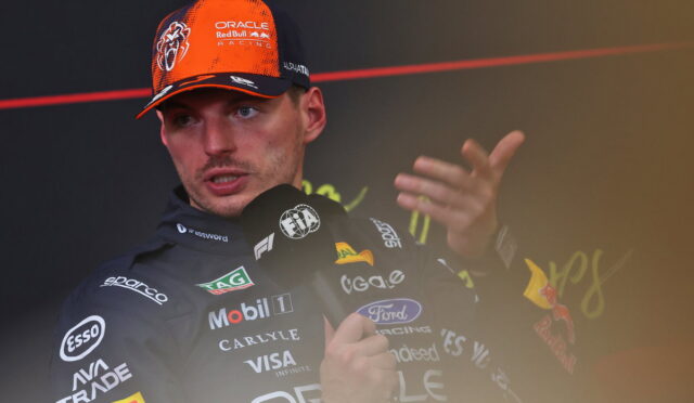 Male race car driver wearing an orange and black Oracle Red Bull Racing cap giving a press conference, speaking into multiple microphones with team logos visible on his suit.