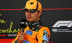F1 driver in an orange suit and cap speaks into a handheld microphone during a post-race interview.