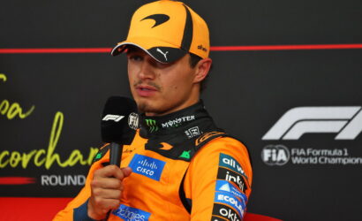 F1 driver in an orange suit and cap speaks into a handheld microphone during a post-race interview.
