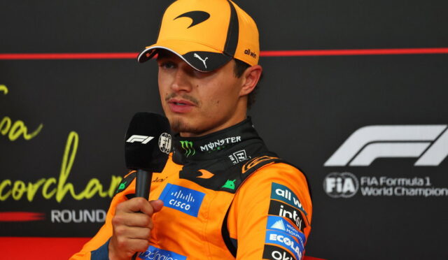 F1 driver in an orange suit and cap speaks into a handheld microphone during a post-race interview.