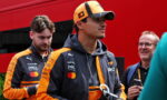Two men in orange and black racing jackets stand outdoors; foreground man wears a cap and holds a water bottle.