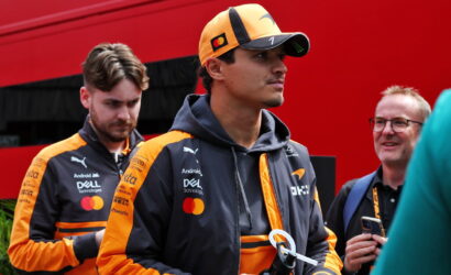 Two men in orange and black racing jackets stand outdoors; foreground man wears a cap and holds a water bottle.