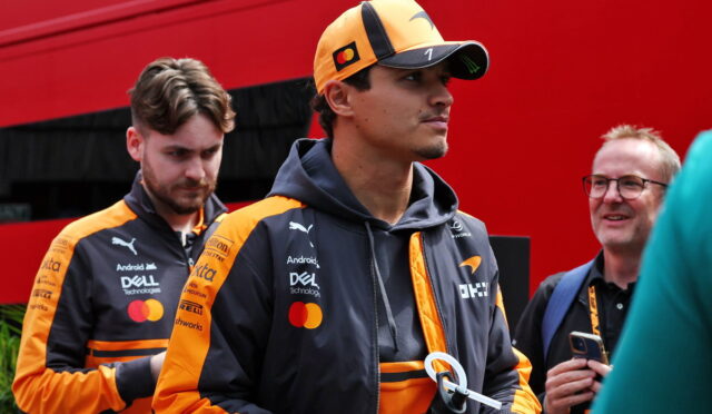 Two men in orange and black racing jackets stand outdoors; foreground man wears a cap and holds a water bottle.