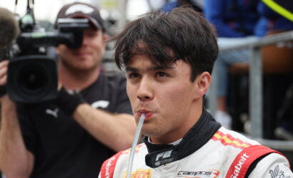 Race car driver in a white and red suit sipping through a straw as a cameraman films in the background