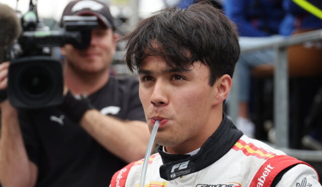 Race car driver in a white and red suit sipping through a straw as a cameraman films in the background