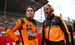 Two race team members in orange suits stand at the pit wall, one driver and the other a crew chief with a headset, looking intently toward the track as a crowd watches behind a fence.