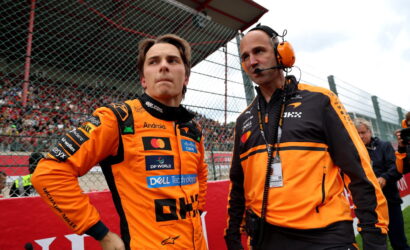 Two race team members in orange suits stand at the pit wall, one driver and the other a crew chief with a headset, looking intently toward the track as a crowd watches behind a fence.