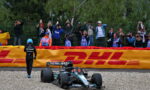 Mercedes Formula 1 car on gravel near a yellow DHL barrier, with a crew member walking nearby as spectators film from behind a fence.