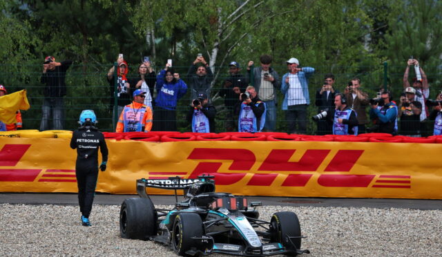 Mercedes Formula 1 car on gravel near a yellow DHL barrier, with a crew member walking nearby as spectators film from behind a fence.