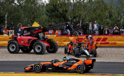Orange Formula 1 car on track as marshals guide a damaged car with a crane-mounted tow tractor behind a yellow barrier; spectators watch.