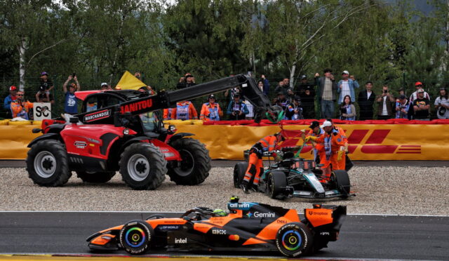 Orange Formula 1 car on track as marshals guide a damaged car with a crane-mounted tow tractor behind a yellow barrier; spectators watch.
