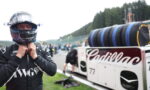 Driver in a black racing suit lowers zipped collar, wearing a full-face helmet, beside a white Cadillac race car with stacked tires in the pit area