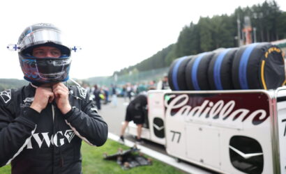 Driver in a black racing suit lowers zipped collar, wearing a full-face helmet, beside a white Cadillac race car with stacked tires in the pit area