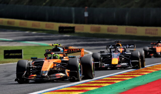 Orange Formula 1 car leading a race with trailing cars on a curvy track and colorful painted runoff areas.