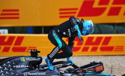 F1 driver in teal and black suit with a blue helmet jumps off a Mercedes-AMG Formula 1 car; DHL orange barrier looms in the background.