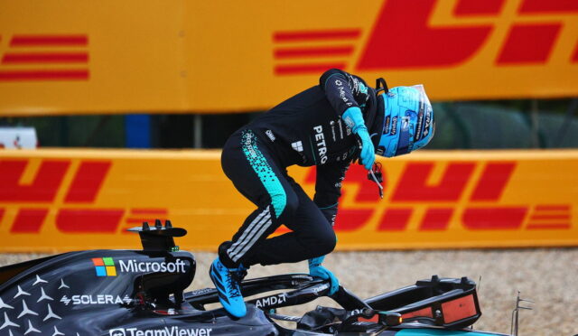 F1 driver in teal and black suit with a blue helmet jumps off a Mercedes-AMG Formula 1 car; DHL orange barrier looms in the background.