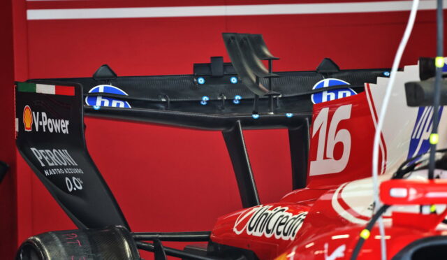 Rear wing of a red Formula 1 car with the number 16 and sponsor logos, in a garage or workshop setting.