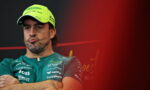 Man in teal sponsor shirt and neon cap with crossed arms, pursed lips, against a dark background with red blur on the right (press event).