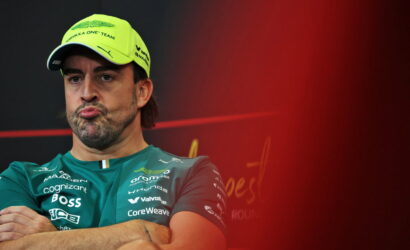 Man in teal sponsor shirt and neon cap with crossed arms, pursed lips, against a dark background with red blur on the right (press event).
