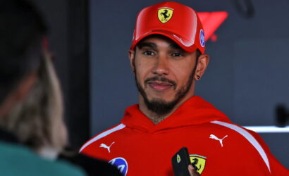 Man in a red Ferrari cap and hoodie being interviewed, holding a phone, with interviewer in foreground left.