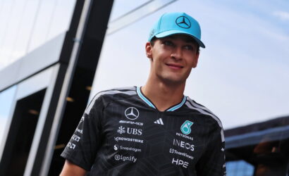 Young man in a Mercedes racing jacket with sponsor logos and a light blue cap, outdoors beside a glass building.