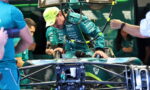 Race car driver in teal suit sits in a Formula 1 car in a pit area, team members assist with the vehicle prep.