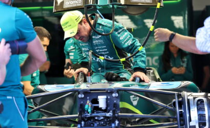 Race car driver in teal suit sits in a Formula 1 car in a pit area, team members assist with the vehicle prep.