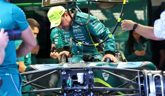 Race car driver in teal suit sits in a Formula 1 car in a pit area, team members assist with the vehicle prep.
