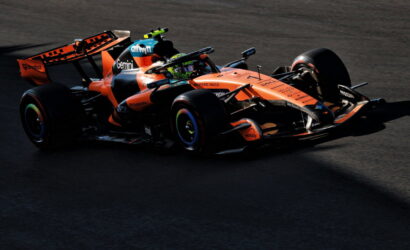 Orange Formula 1 car on a dark track, branding and sponsor logos visible as it corners.