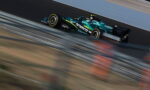 A teal and black Formula-style race car speeding along a track with motion blur foreground and background barriers nearby