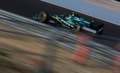 A teal and black Formula-style race car speeding along a track with motion blur foreground and background barriers nearby
