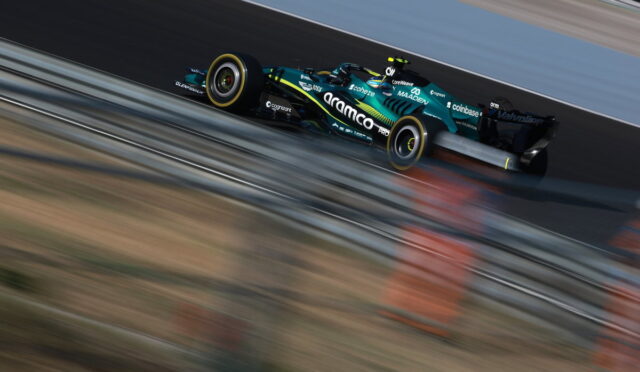 A teal and black Formula-style race car speeding along a track with motion blur foreground and background barriers nearby