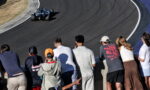 Blue open-wheel race car on a curved asphalt track, viewed from the spectator wall where a group of fans watches intently.