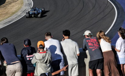 Blue open-wheel race car on a curved asphalt track, viewed from the spectator wall where a group of fans watches intently.
