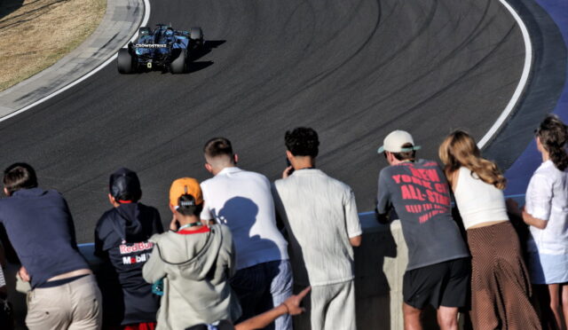 Blue open-wheel race car on a curved asphalt track, viewed from the spectator wall where a group of fans watches intently.