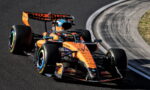 Orange McLaren Formula 1 car on a race track mid-turn, with curved barrier nearby.