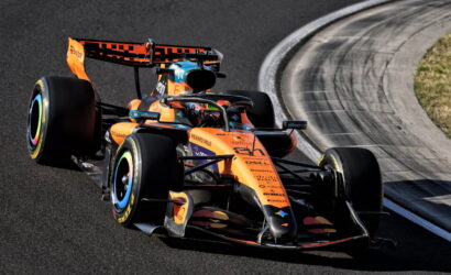 Orange McLaren Formula 1 car on a race track mid-turn, with curved barrier nearby.