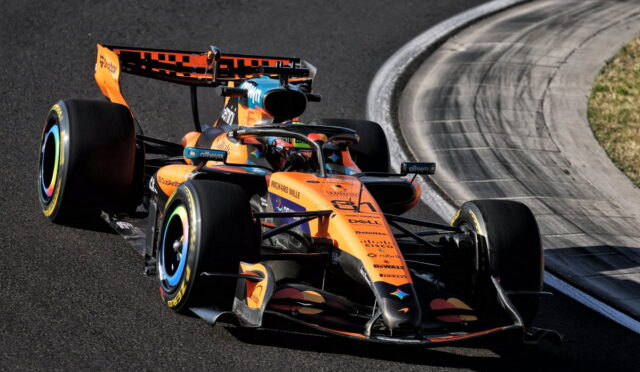 Orange McLaren Formula 1 car on a race track mid-turn, with curved barrier nearby.