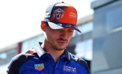 Man wearing an Oracle Red Bull Racing cap and blue team polo, looking down at a racing facility exterior behind him.